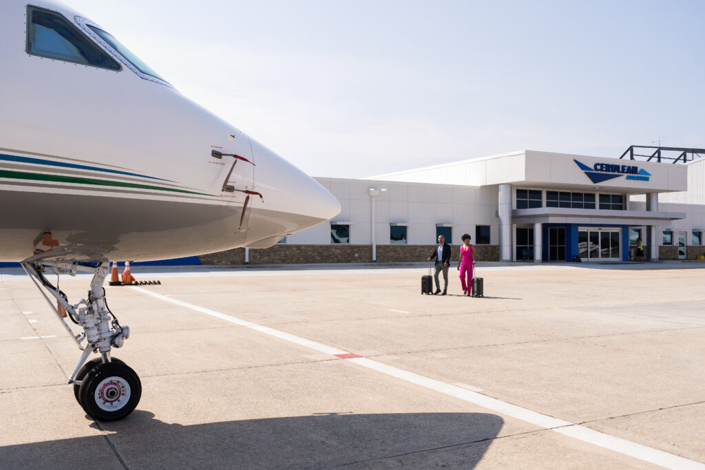 A private jet is parked on the tarmac in front of the Cerulean Aviation terminal building. In the background, a man and woman, both dressed in business attire, walk toward the aircraft, each pulling a suitcase.