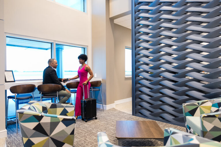 A man and woman greet each other with a handshake in a modern airport lounge. The woman, holding a rolling suitcase, stands near the man who is seated at a counter with barstools by large windows overlooking the airfield. The lounge features contemporary decor, including patterned chairs and a woven design partition.