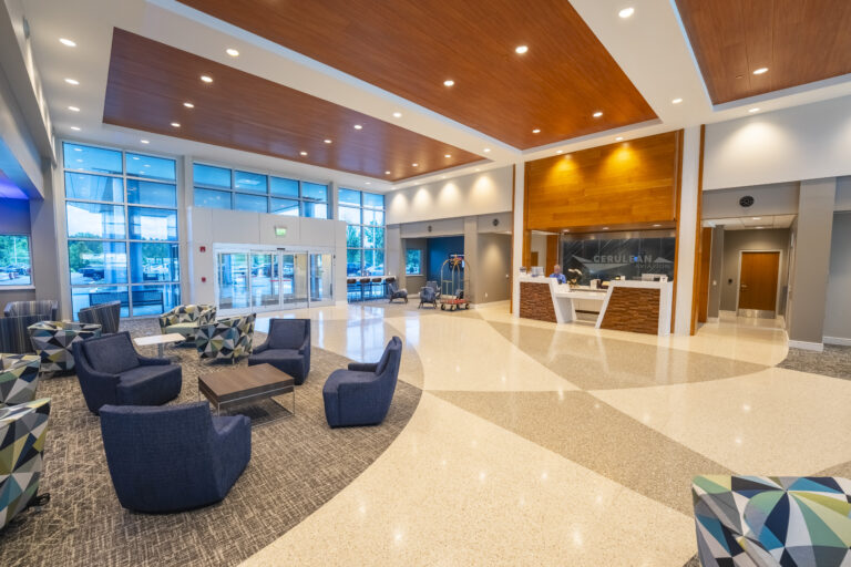 A spacious and modern airport lobby at Cerulean Aviation, featuring a reception desk staffed by two employees, comfortable seating areas with patterned and blue chairs, a polished terrazzo floor, and a wood-accented ceiling. Large windows provide natural light and views of the exterior.
